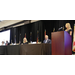 A group of people sitting on a panel with a woman speaking at a podium