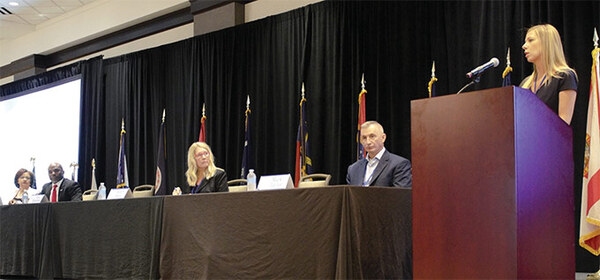 A group of people sitting on a panel with a woman speaking at a podium