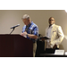 a man speaking at a podium referencing his notes while another man stands beside him waiting to present