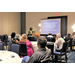 A woman standing in front of a screen with a PowerPoint showing while speaking to a room full of people seated at tables 