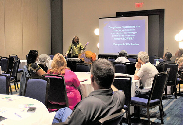 A woman standing in front of a screen with a PowerPoint showing while speaking to a room full of people seated at tables 