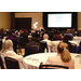 A room full of people listening to a presentation while seated at round tables