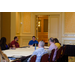 Men and women are seated around a table and they listen as the man at the head of the table is speaking.