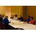 Men and women take notes and listen as they are gathered around the table. 