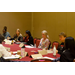 Women and men are seated around a table listening to a speaker and taking notes.