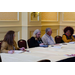 Three women and a man sit at a table. Two of the women are taking notes while the other man and woman talk.