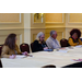 Three women and a man sit at a table. Two of the women are take notes and the woman and man talk.