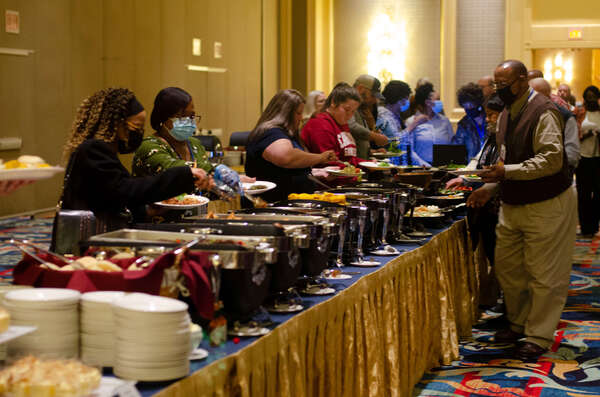 Catering dishes full of food line the table as people fill their plates.