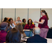 A woman wearing glasses stands to the right speaking to those who are seated at tables around her.