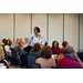 A woman stands in the middle of a room with people seated at tables around her. 