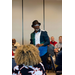 A man wearing glasses and a hat, stands in a room full of seated people. 