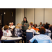 A woman stands in the middle of tables of people and speaks using hand motions.