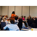 A woman is speaking to tables of people using hand motions.