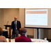 A man stands to the left of a presentation he is giving and speaks using hand motions.