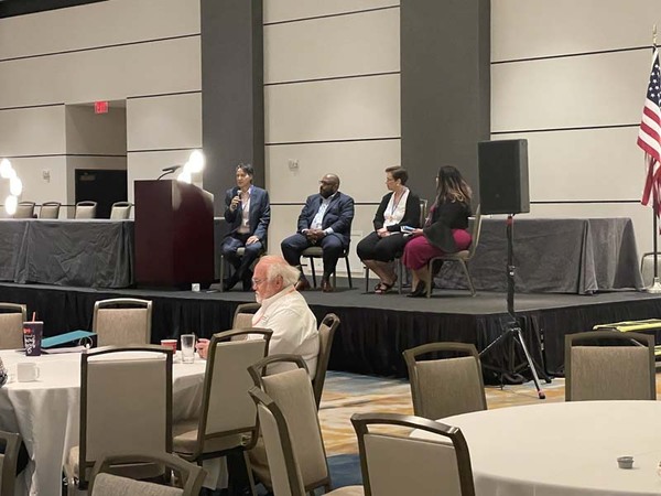  A panel of four speakers are sitting on a stage in a large conference room. 