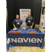 Two men stand with their hands in their pockets behind a booth table for Navien. 