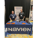 Two men stand with their hands in their pockets behind a booth table for Navien.