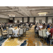 A conference room full of people at multiple tables listening to a person on stage speak. 