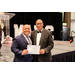  Two men stand and smile by the state holding an award. 