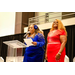  Two women stand on the stage, one of them talking at the podium. 