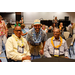  Two men sitting at a table with a third man standing between them. All of them are wearing colorful leis. 