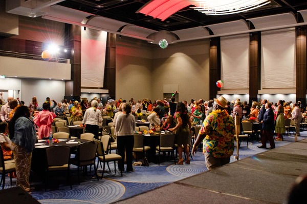 A wide shot of a crowded room standing and hitting three beach balls back and forth.