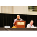  Anthony Goodson speaking at the podium, while Mike Sweet sits next to him. 