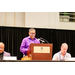  A man standing and speaking at the podium while President Anthony Goodson, and Mike Sweet sit on either side. 