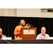  A woman standing and speaking at the podium while President Anthony Goodson, and Mike Sweet sit on either side. 