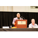  President Anthony Goodson speaking at the podium with Mike Sweet sitting to his left. 