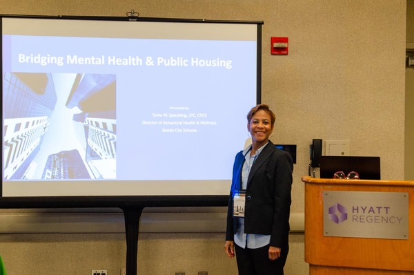 Tonia W. Spaulding, LPC, CPCS, Director of Behavioral Health and Wellness at Dublin City School smiling as she stands in front of her Powerpoint about Bridging Mental Health and Public Housing.