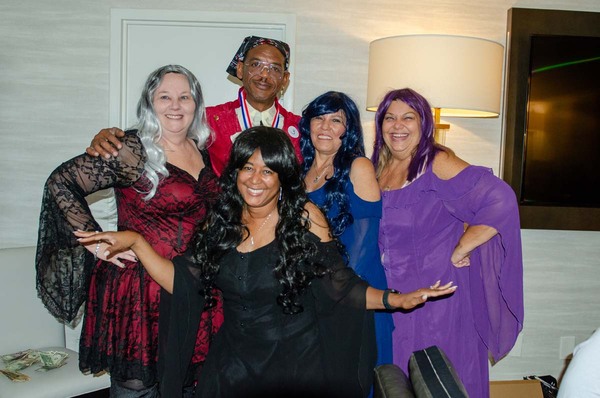 Four women in various colorful dresses posing with a man in a double breasted brocade waistcoat.